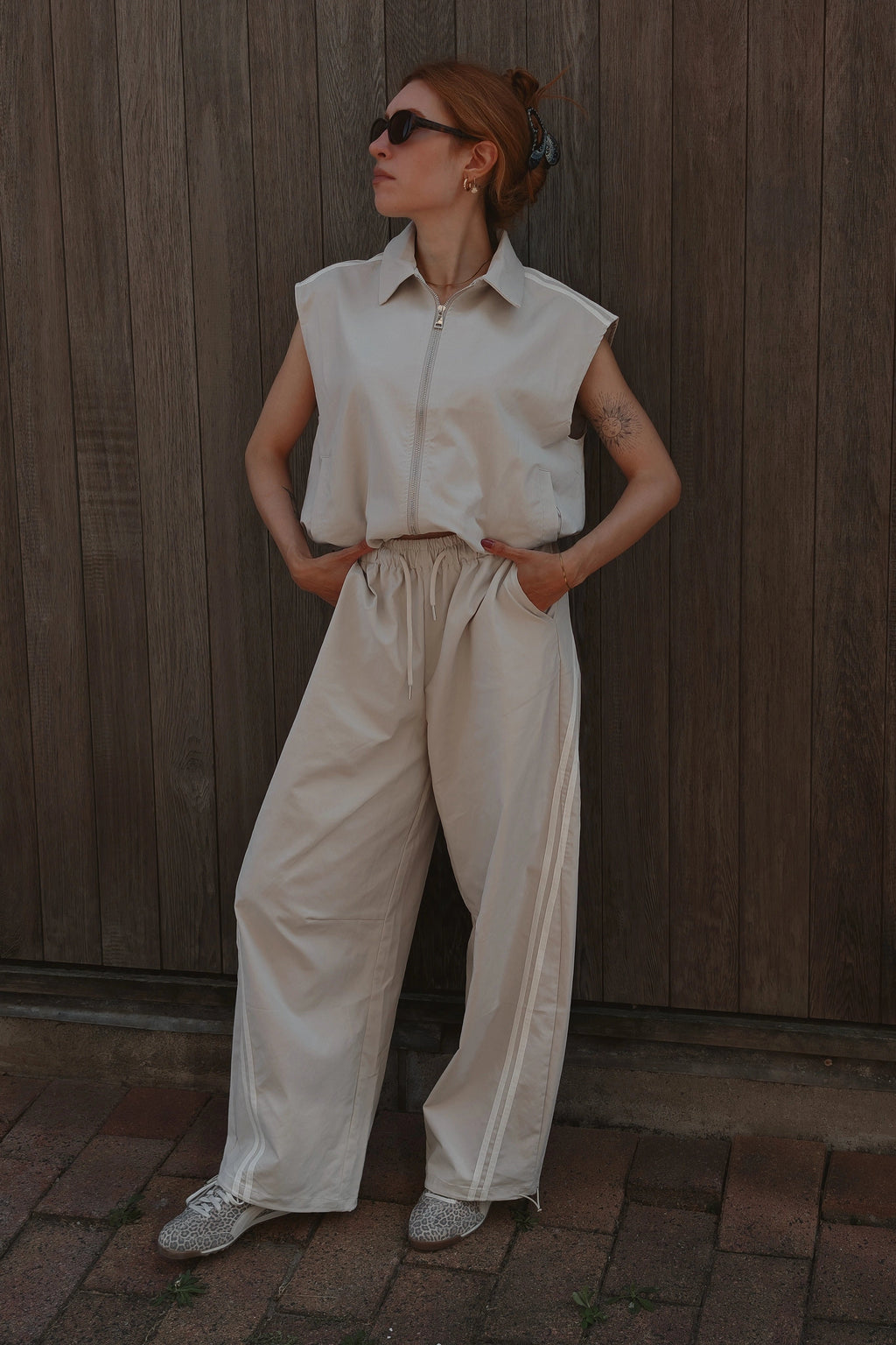 A woman wearing a sleeveless cream-colored collared shirt and matching wide-leg trousers, standing against a dark wooden plank wall.