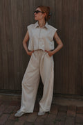 A woman wearing a sleeveless cream-colored collared shirt and matching wide-leg trousers, standing against a dark wooden plank wall.