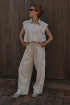 A woman wearing a sleeveless cream-colored collared shirt and matching wide-leg trousers, standing against a dark wooden plank wall.