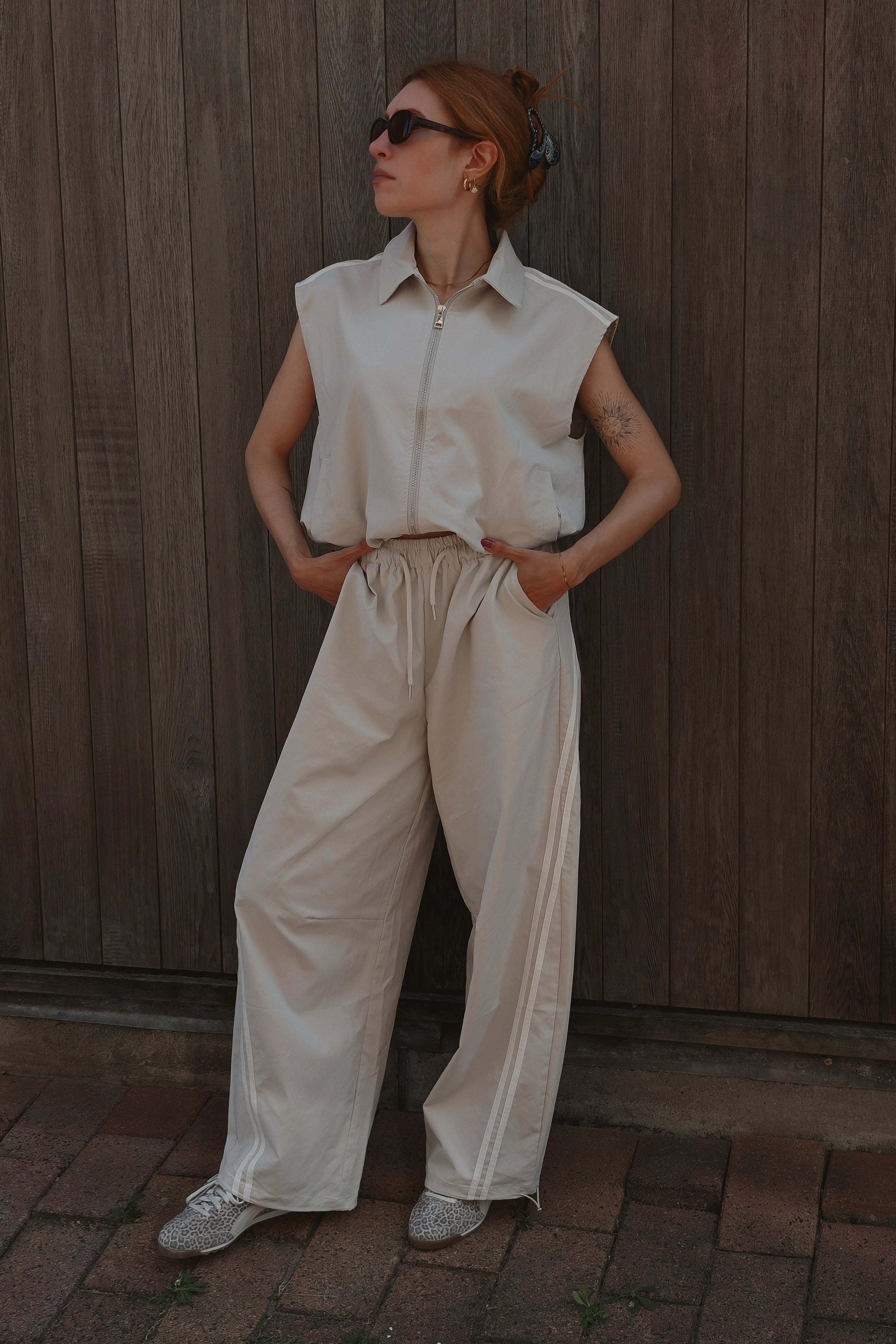 A woman wearing a sleeveless cream-colored collared shirt and matching wide-leg trousers, standing against a dark wooden plank wall.