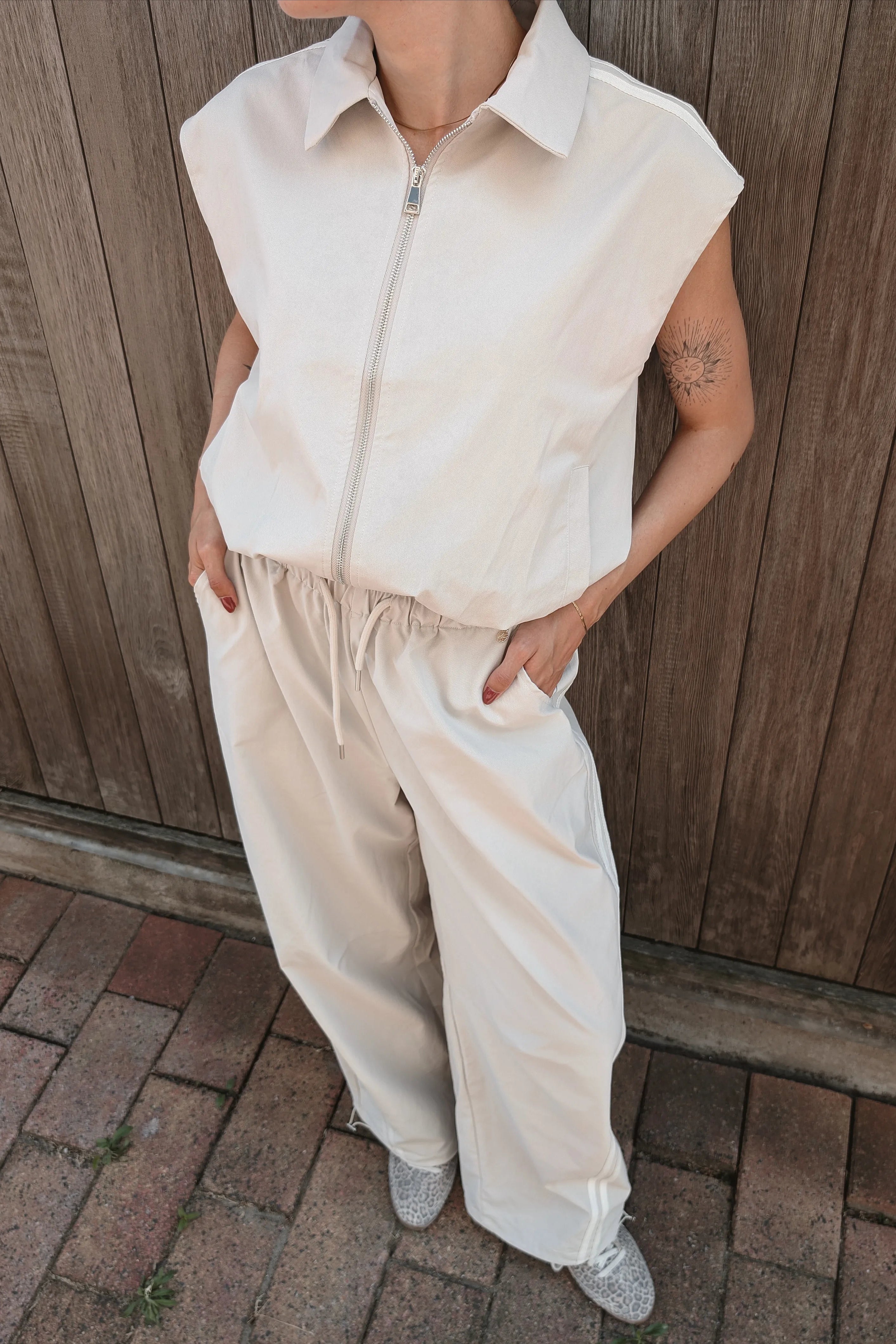 A person wearing a sleeveless zip-front collared top and matching wide-leg drawstring pants, standing outdoors against a wooden fence with brick pavers below.