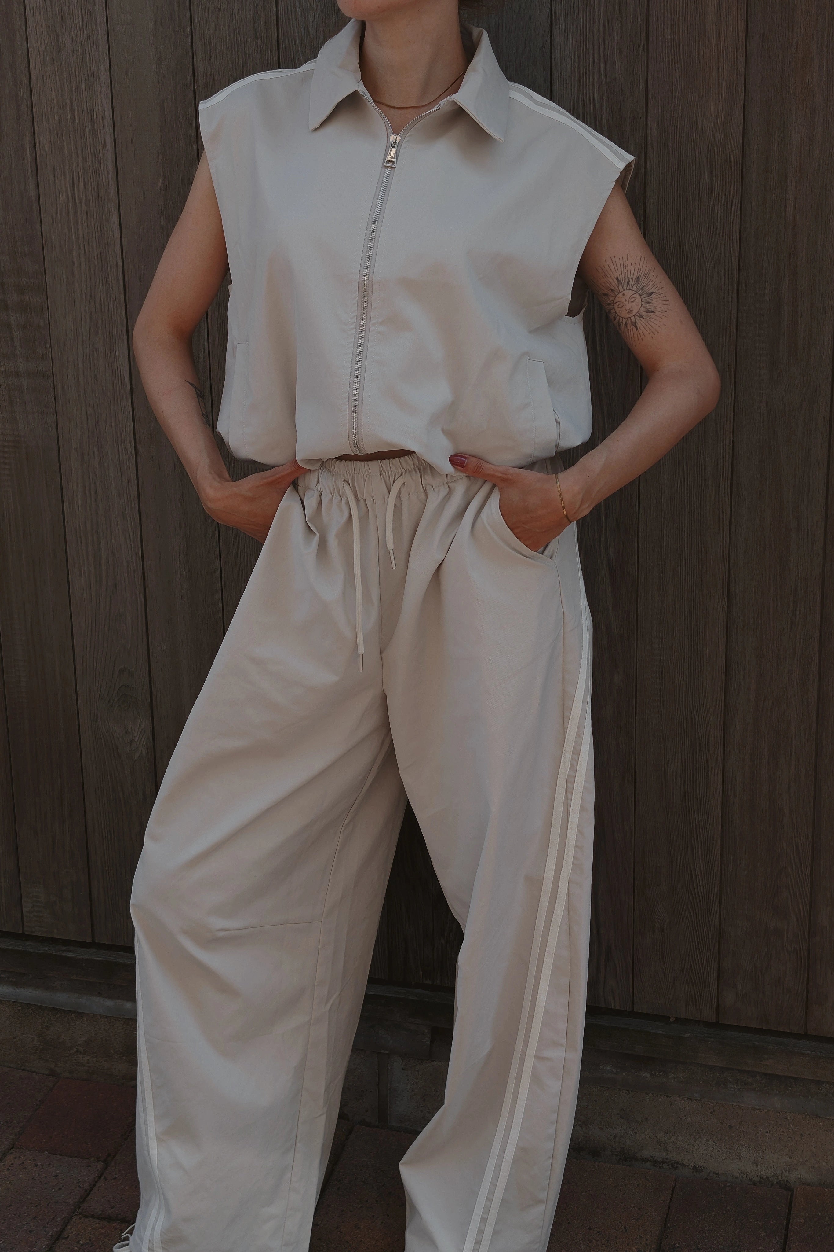 A person wearing a sleeveless collared zip-front top and matching wide-leg drawstring pants, standing against a dark wooden plank wall.