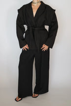A model wearing a black belted wrap jacket and matching wide-leg trousers, styled with black sandals and a silver choker necklace against a plain light gray background.