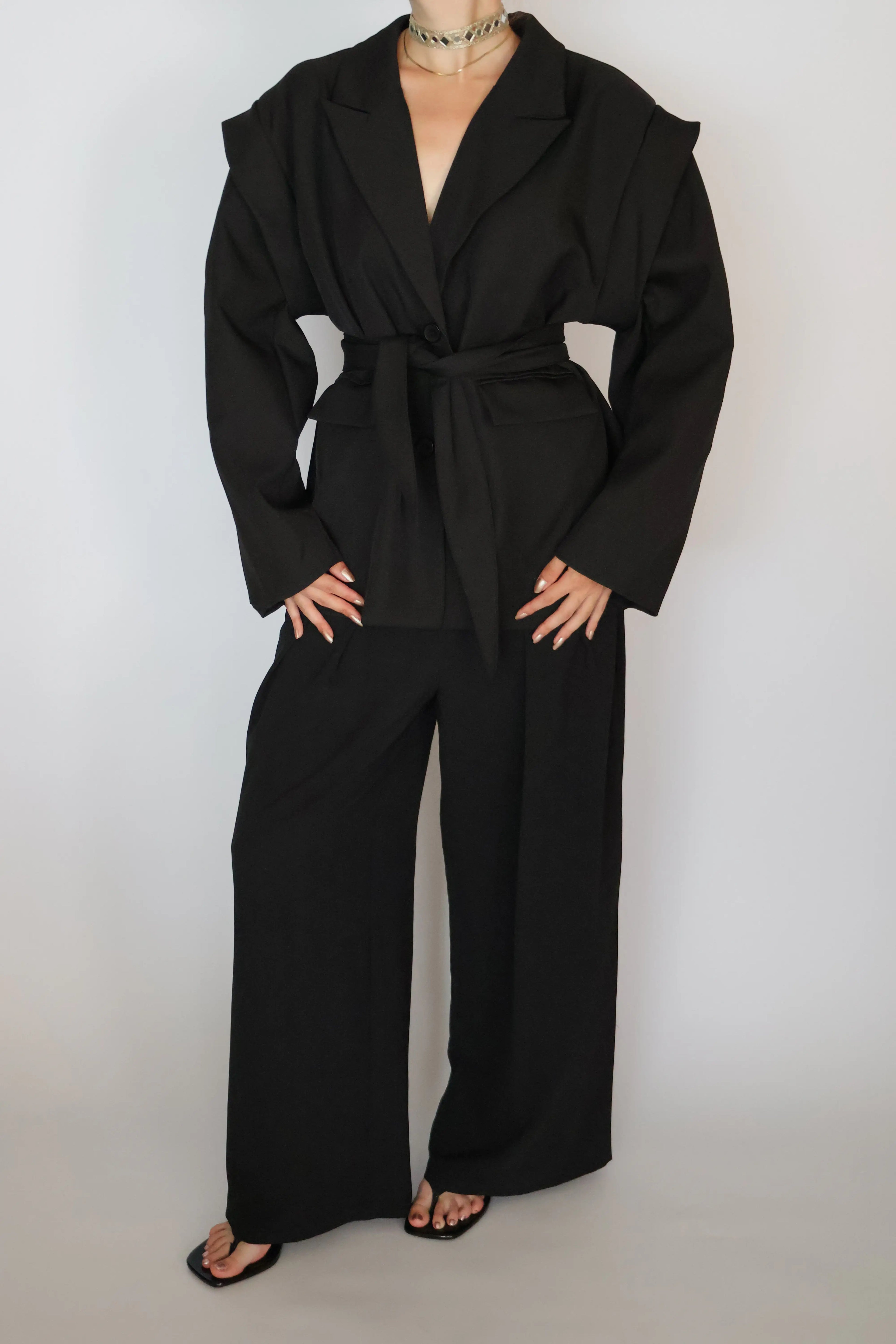 A model wearing a black belted wrap jacket and matching wide-leg trousers, styled with black sandals and a silver choker necklace against a plain light gray background.
