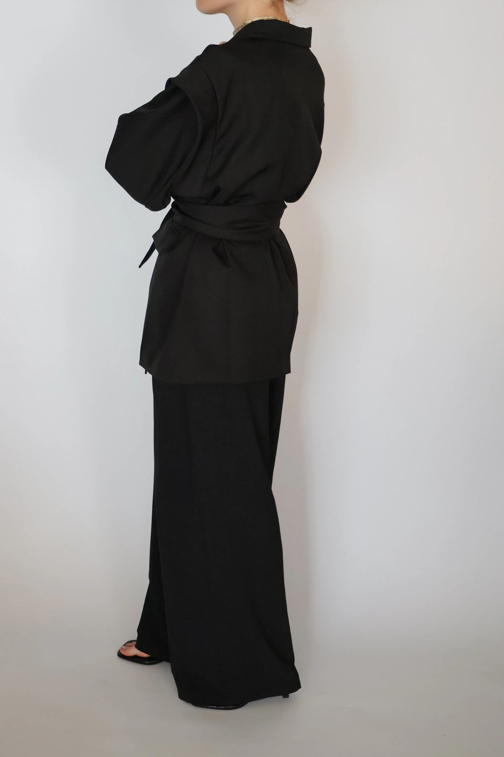 A model shown from the back wearing a black belted long-sleeve top and matching wide-leg trousers against a plain light gray background.
