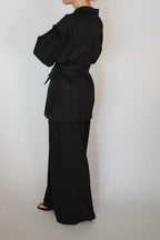 A model shown from the back wearing a black belted long-sleeve top and matching wide-leg trousers against a plain light gray background.