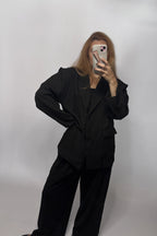A woman takes a mirror selfie wearing a black oversized blazer and matching wide-leg trousers against a plain light gray studio backdrop.