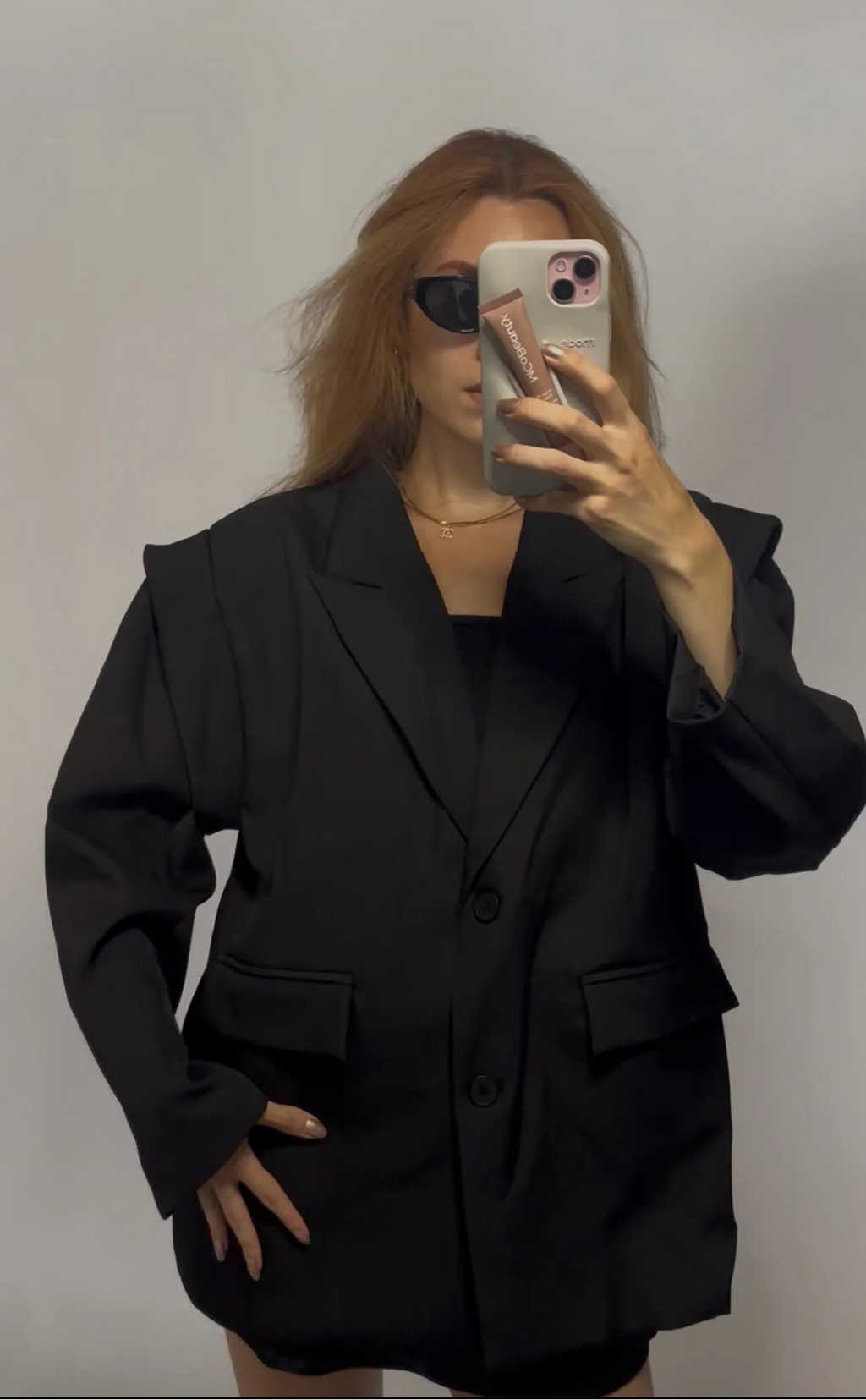 A woman takes a mirror selfie wearing an oversized black blazer with structured shoulders and flap pockets, paired with a black top and black shorts, against a plain light gray background.
