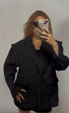 A woman takes a mirror selfie wearing an oversized black blazer with structured shoulders and flap pockets, paired with a black top and black shorts, against a plain light gray background.