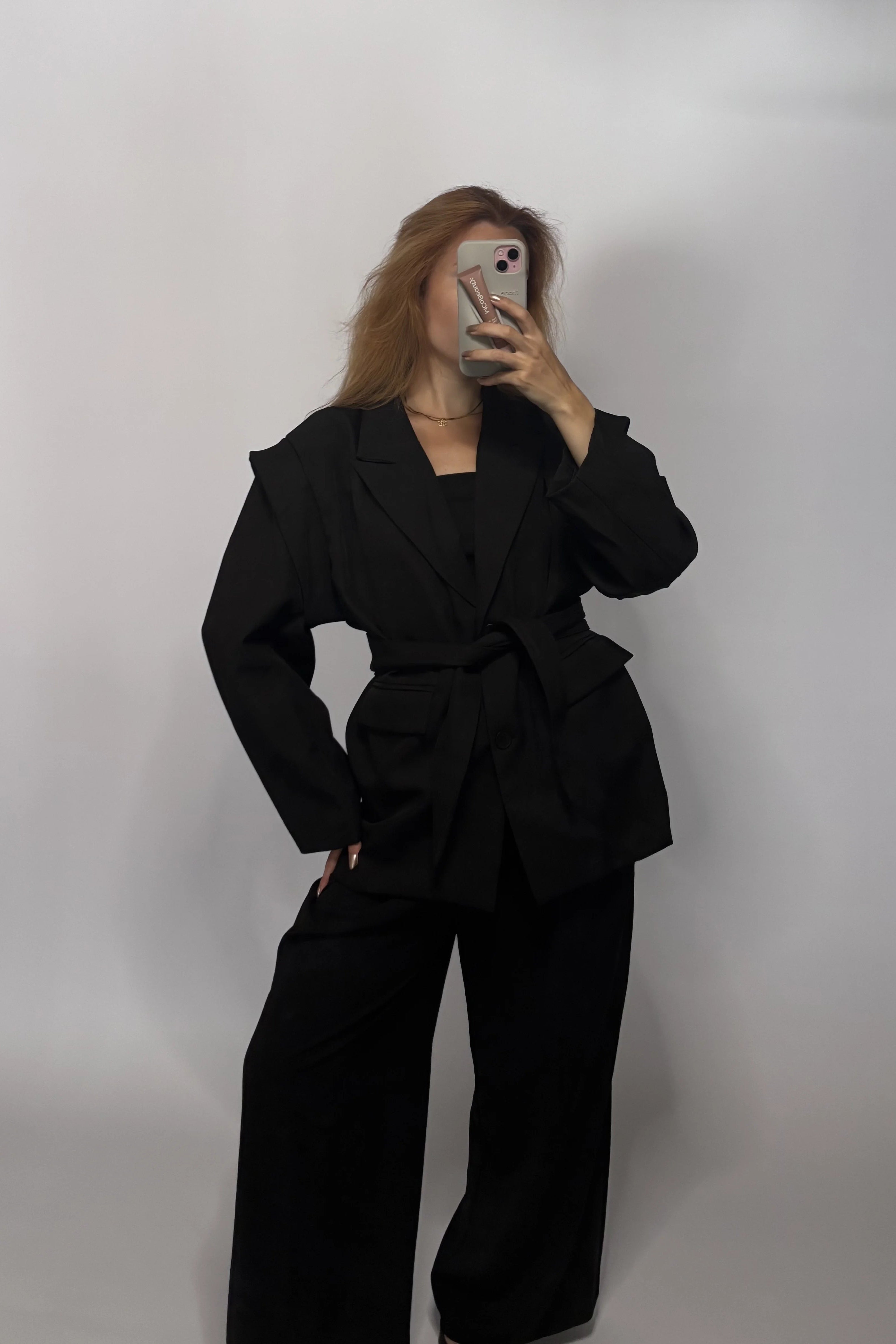 A woman takes a mirror selfie wearing a black belted blazer and matching wide-leg trousers against a plain light gray background.