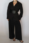 A model wearing a black long-sleeve wrap-style blazer with a wide black faux-leather belt, paired with black wide-leg trousers and black strappy heels, against a plain light gray background.