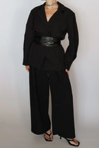 A model wearing a black long-sleeve wrap-style blazer with a wide black faux-leather belt, paired with black wide-leg trousers and black strappy heels, against a plain light gray background.