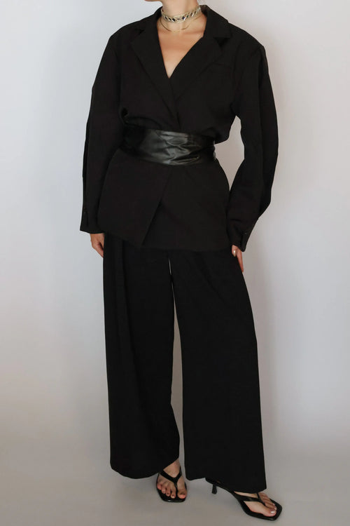 A model wearing a black long-sleeve wrap-style blazer with a wide black faux-leather belt, paired with black wide-leg trousers and black strappy heels, against a plain light gray background.