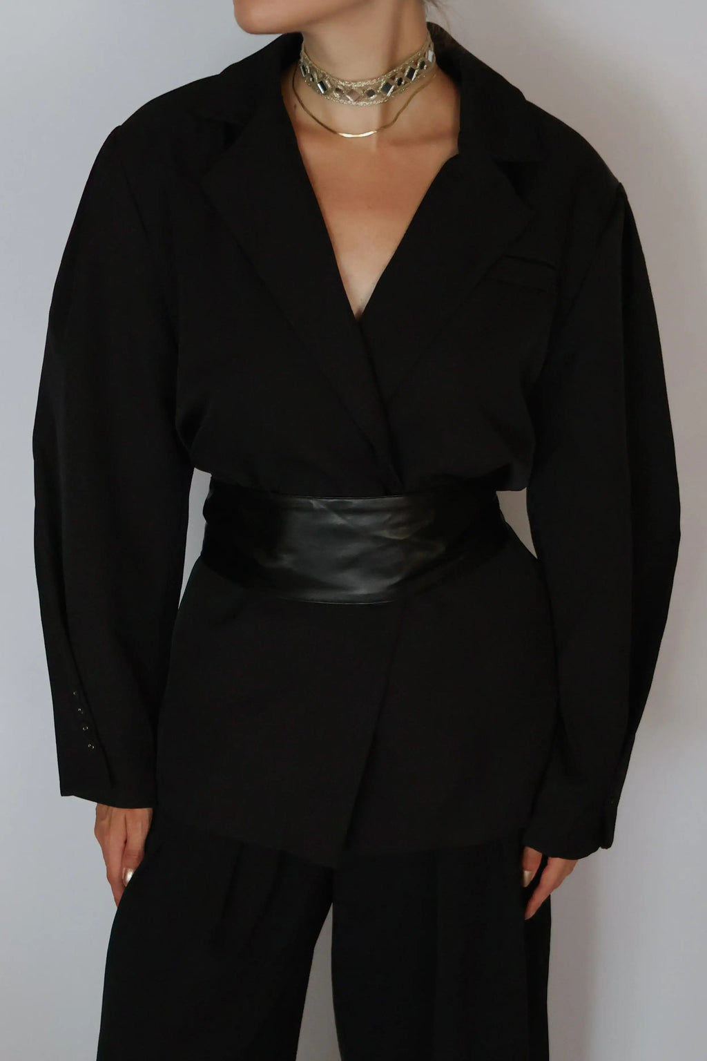 A person wearing a black wrap-style blazer with long sleeves, black trousers, and a wide black faux-leather belt, styled with layered gold chain necklaces against a plain light gray background.