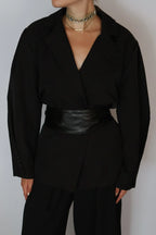 A person wearing a black wrap-style blazer with long sleeves, black trousers, and a wide black faux-leather belt, styled with layered gold chain necklaces against a plain light gray background.