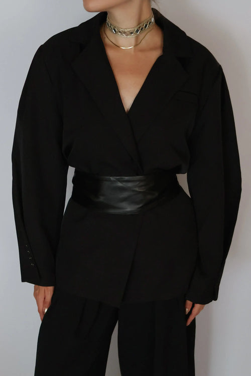 A person wearing a black wrap-style blazer with long sleeves, black trousers, and a wide black faux-leather belt, styled with layered gold chain necklaces against a plain light gray background.