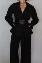 A person wearing a black long-sleeve wrap-style blazer with a wide collar, matching black wide-leg pants, and a shiny black belt at the waist, photographed against a plain light gray wall.