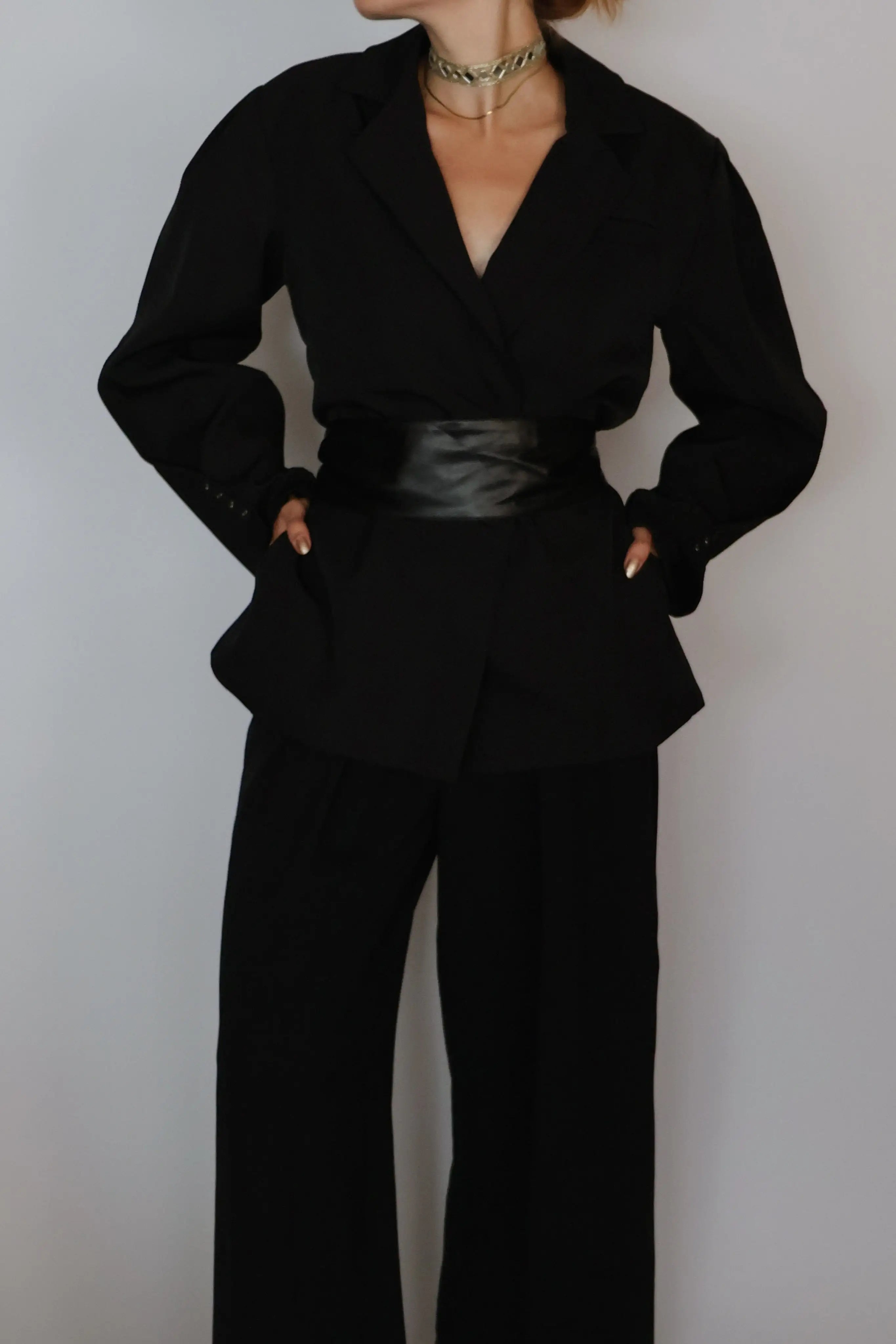 A person wearing a black long-sleeve wrap-style blazer with a wide collar, matching black wide-leg pants, and a shiny black belt at the waist, photographed against a plain light gray wall.