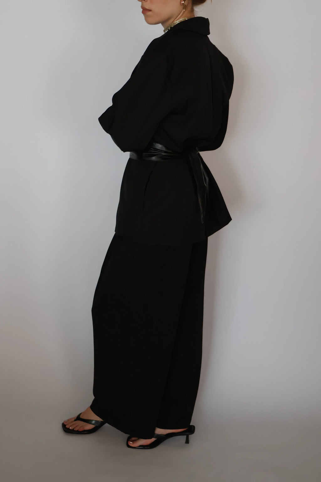 A model shown from the side wearing a black long-sleeve collared top with a matching black belt and wide-leg black trousers, paired with black heeled sandals, against a plain light gray background.