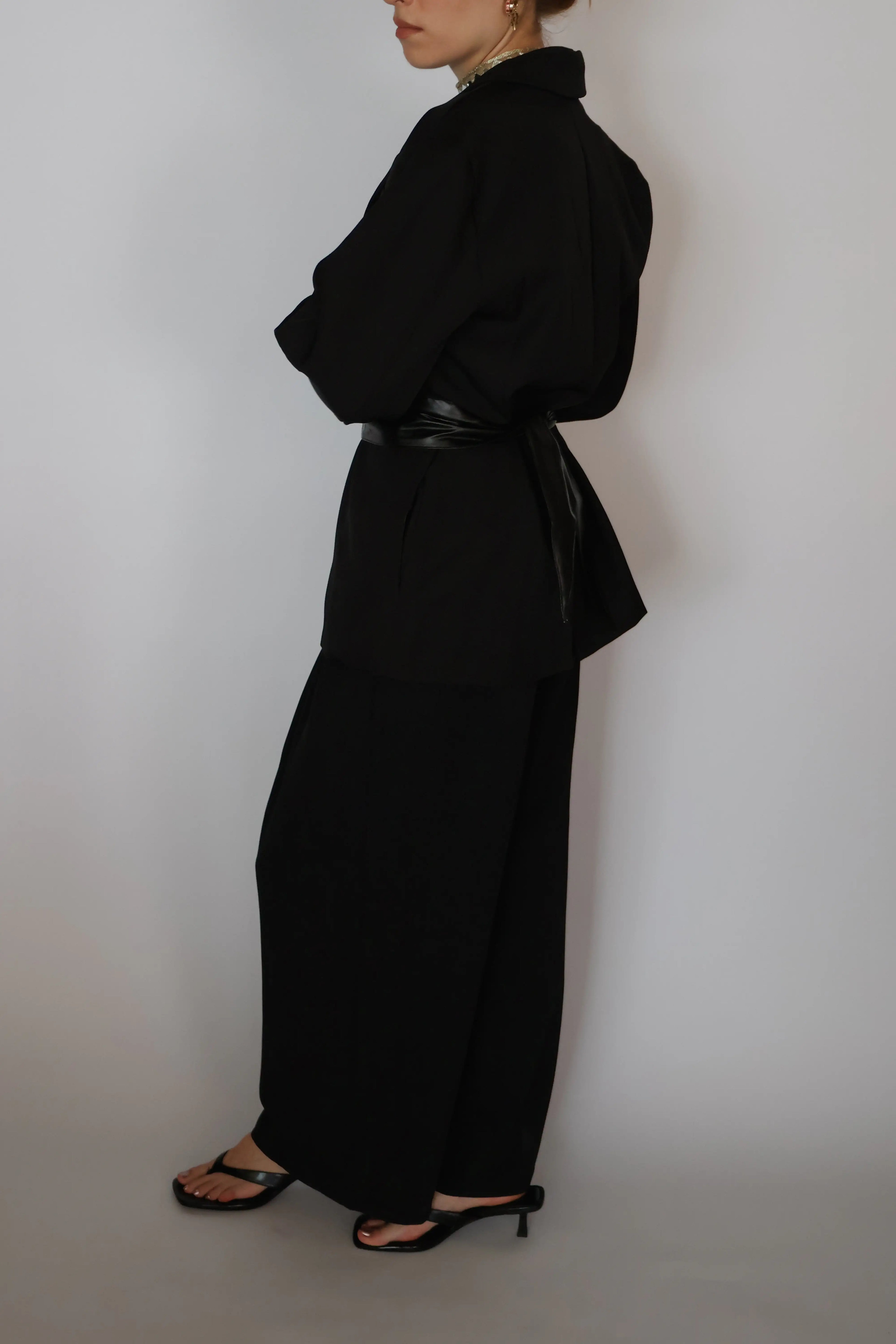 A model shown from the side wearing a black long-sleeve collared top with a matching black belt and wide-leg black trousers, paired with black heeled sandals, against a plain light gray background.