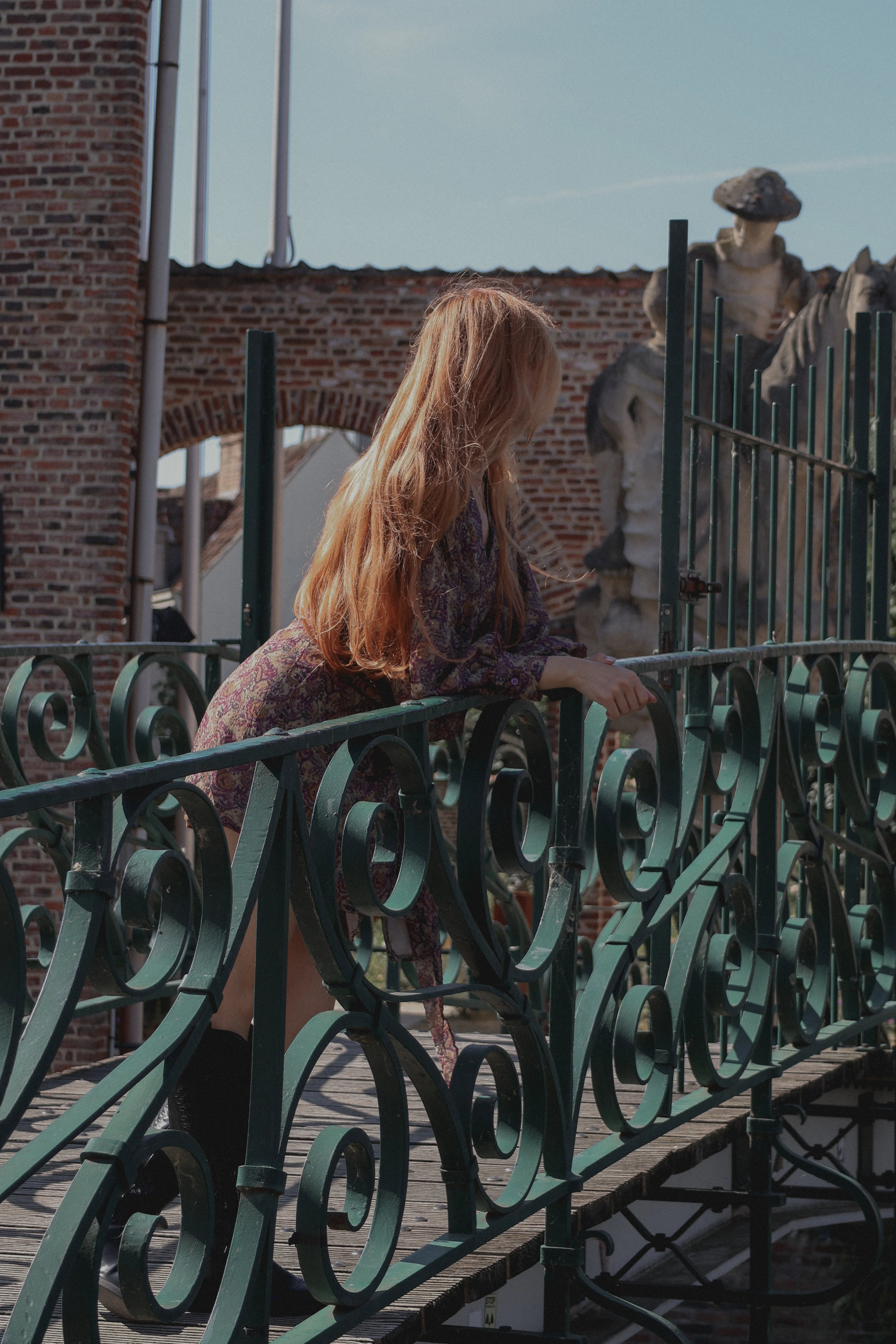 A woman with long strawberry-blonde hair stands on a wooden bridge behind an ornate green metal railing, wearing a purple patterned dress. The background features a brick wall, a stone statue, and a clear blue sky.