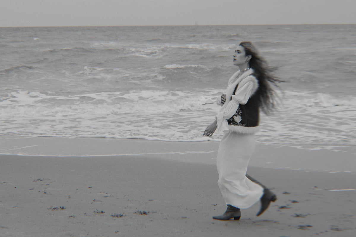 A woman walks along a sandy beach with the ocean waves in the background, wearing a white long-sleeve top, a black vest, and white wide-leg pants.