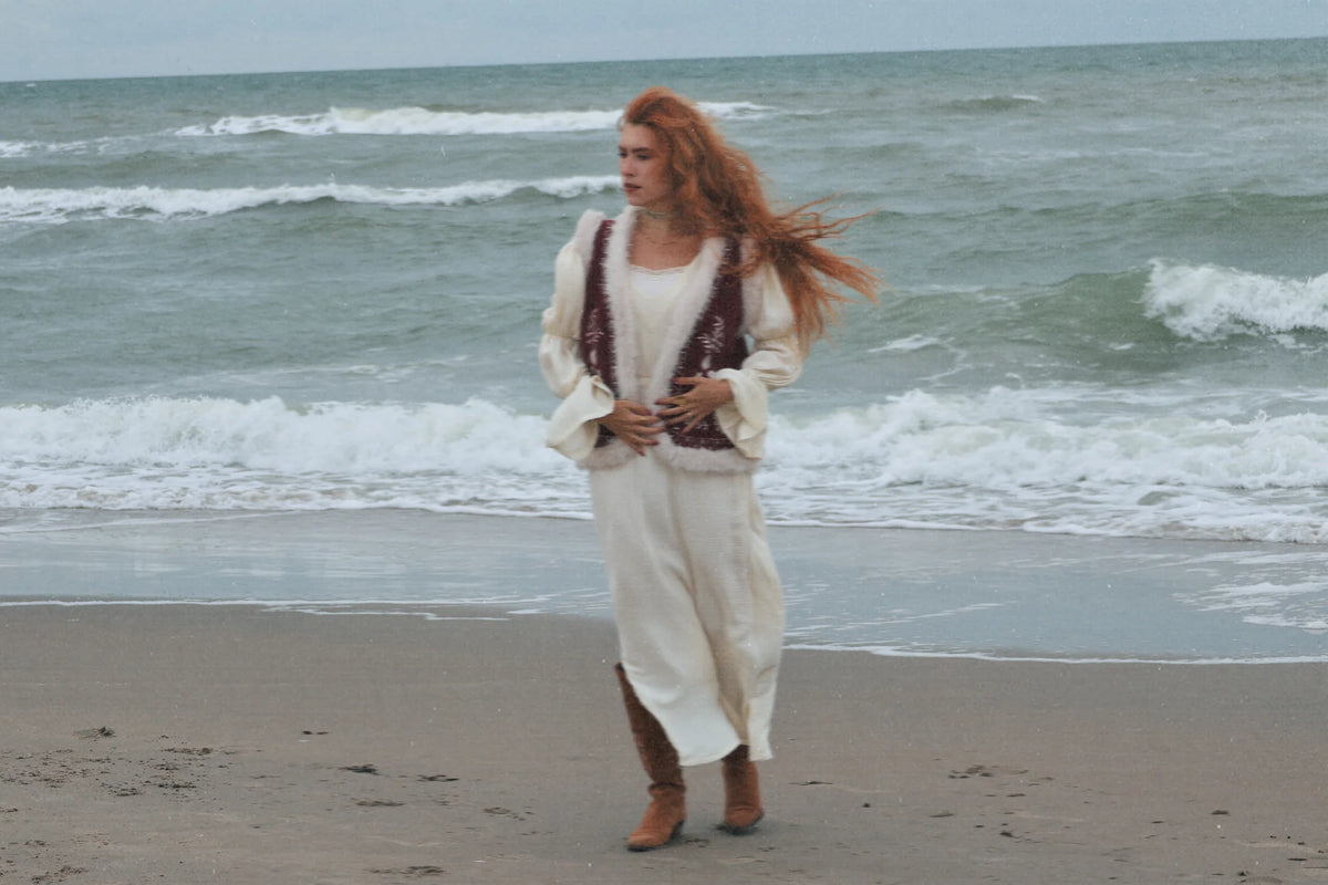 A woman with long red hair stands on a sandy beach with the ocean waves behind her, wearing a loose white dress with dark brown trim and boots.
