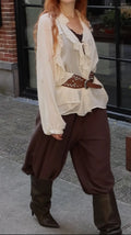 A woman wearing a bohemian-style outfit featuring a cream ruffled long-sleeve blouse, a brown studded belt, loose brown harem-style pants, and dark brown knee-high boots, standing outdoors in front of a building.
