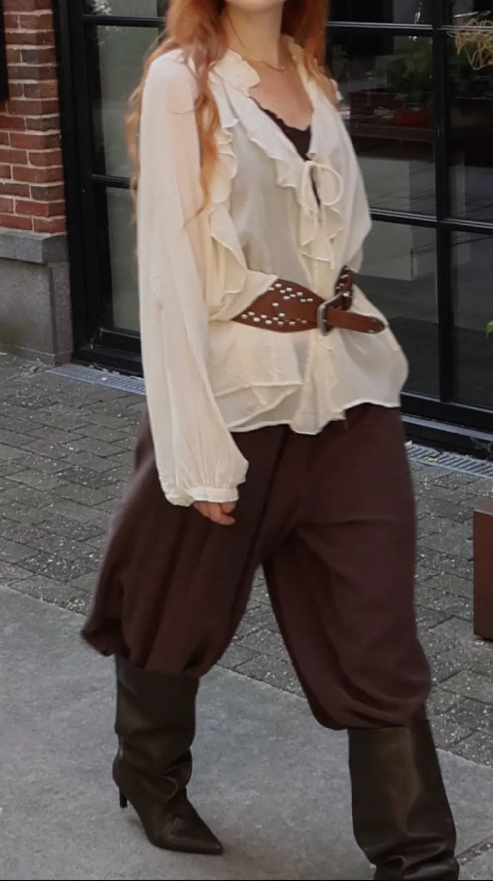 A woman wearing a bohemian-style outfit featuring a cream ruffled long-sleeve blouse, a brown studded belt, loose brown harem-style pants, and dark brown knee-high boots, standing outdoors in front of a building.