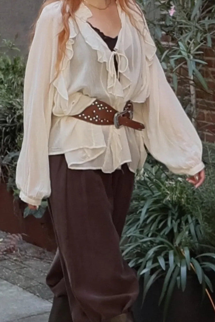 A woman wearing a cream ruffled blouse, dark brown wide-leg pants, a brown studded belt, and dark brown knee-high boots, walking outdoors on a sidewalk next to potted plants.