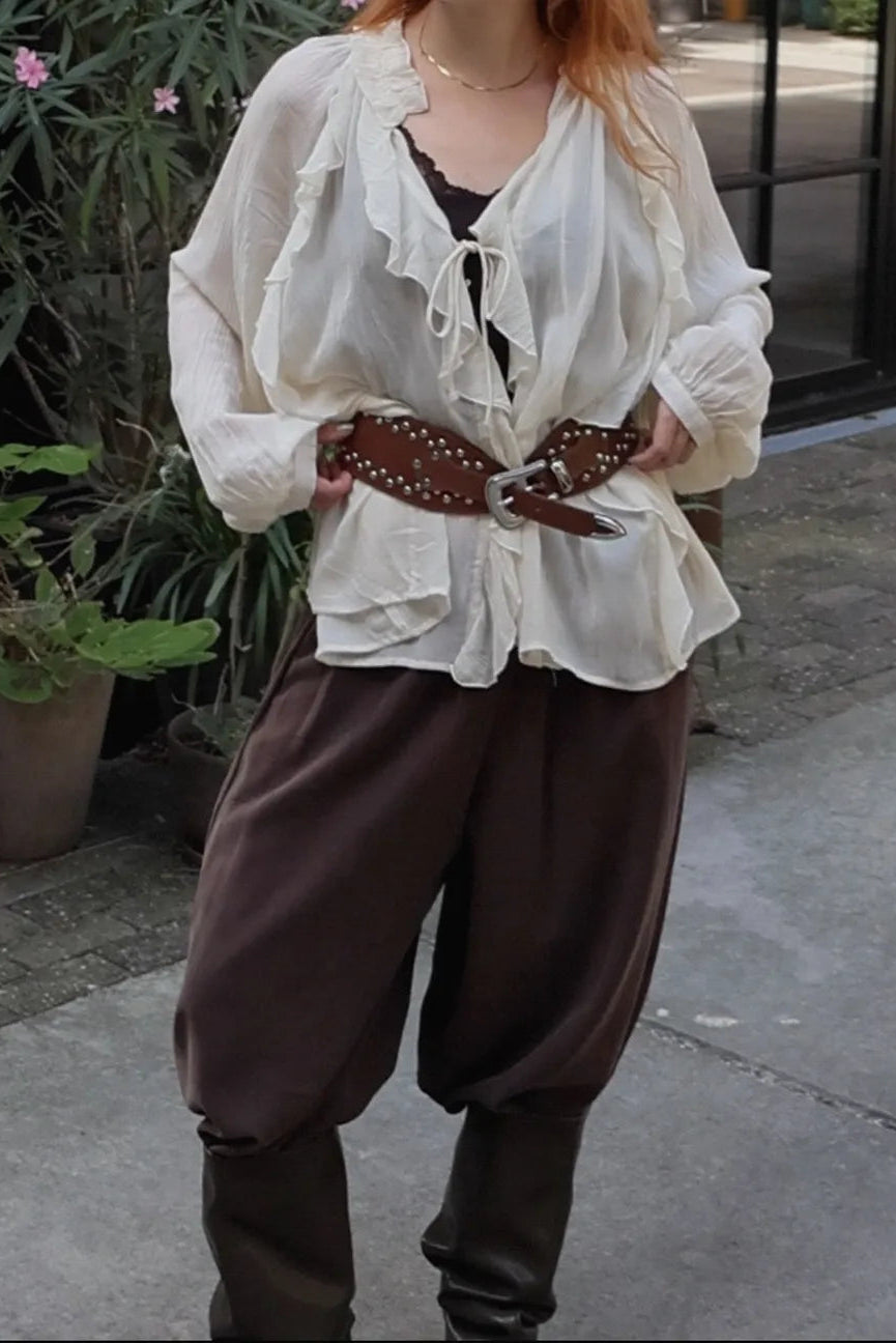 A person wearing a cream ruffled long-sleeve blouse, brown harem-style pants, and a brown studded belt, standing outdoors on a paved walkway with plants in the background.