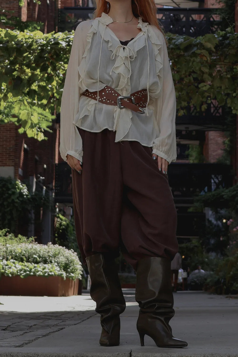 A person standing outdoors wearing a cream ruffled blouse with a brown studded belt, dark brown baggy pants, and dark brown heeled boots.