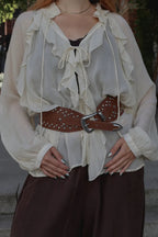 A woman wearing a cream ruffled long-sleeve blouse with a tie neckline, styled with a brown studded belt and dark brown pants.