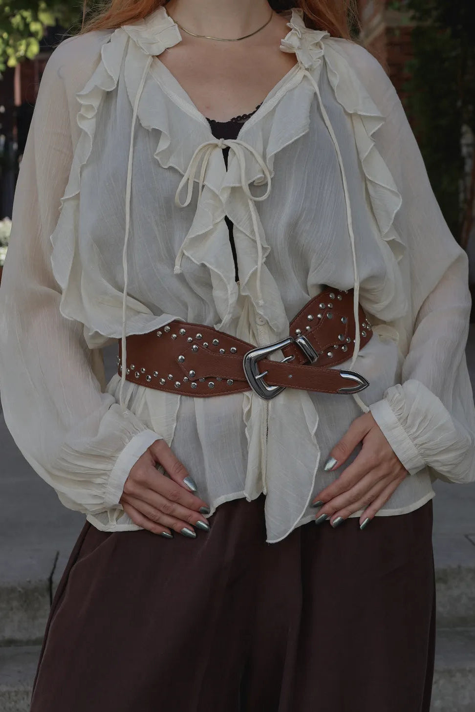 A woman wearing a cream ruffled long-sleeve blouse with a tie neckline, styled with a brown studded belt and dark brown pants.