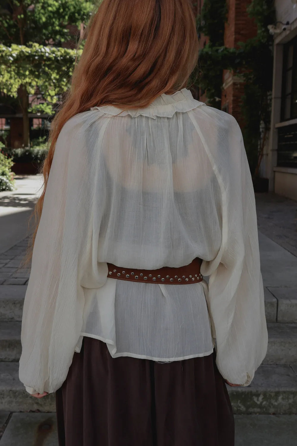 A woman viewed from behind wearing a sheer cream-colored long-sleeve blouse with a ruffled collar, tucked into a white undershirt, a brown studded belt, and dark brown pants.