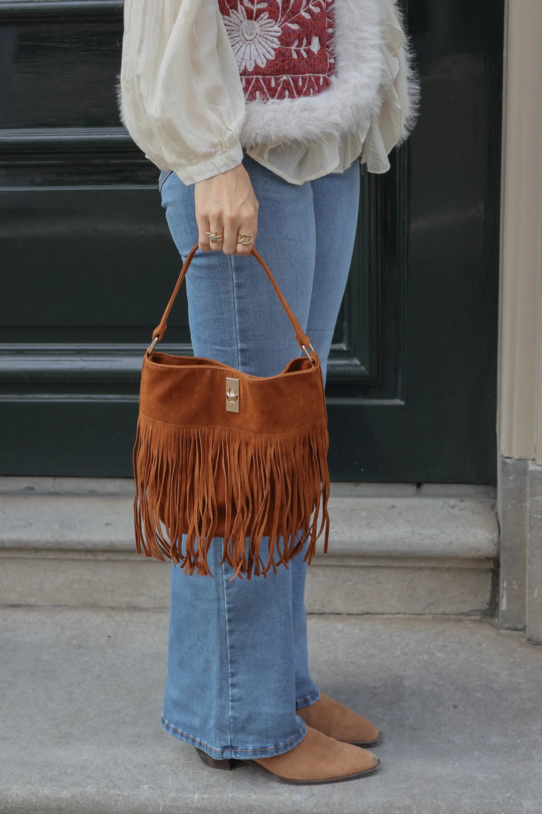 A person wearing blue flared jeans and tan boots holds a brown suede fringe bucket bag in front of a dark door.