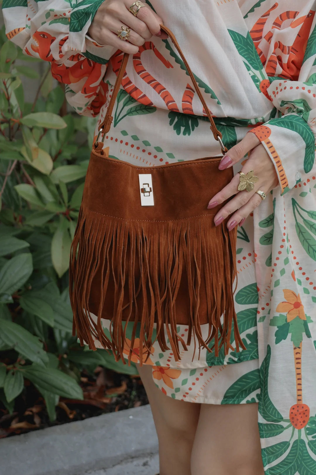 A person wearing a cream floral dress holds a brown suede fringe shoulder bag with a top handle, standing outdoors in front of green plants.