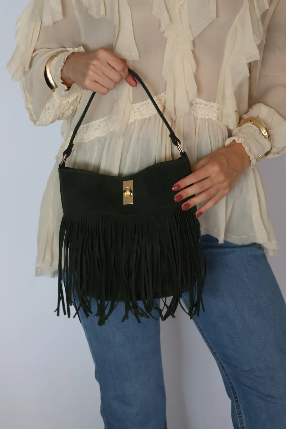 A person wearing a cream ruffled blouse and blue jeans holds a black fringe shoulder bag with a small gold clasp against a plain light background.