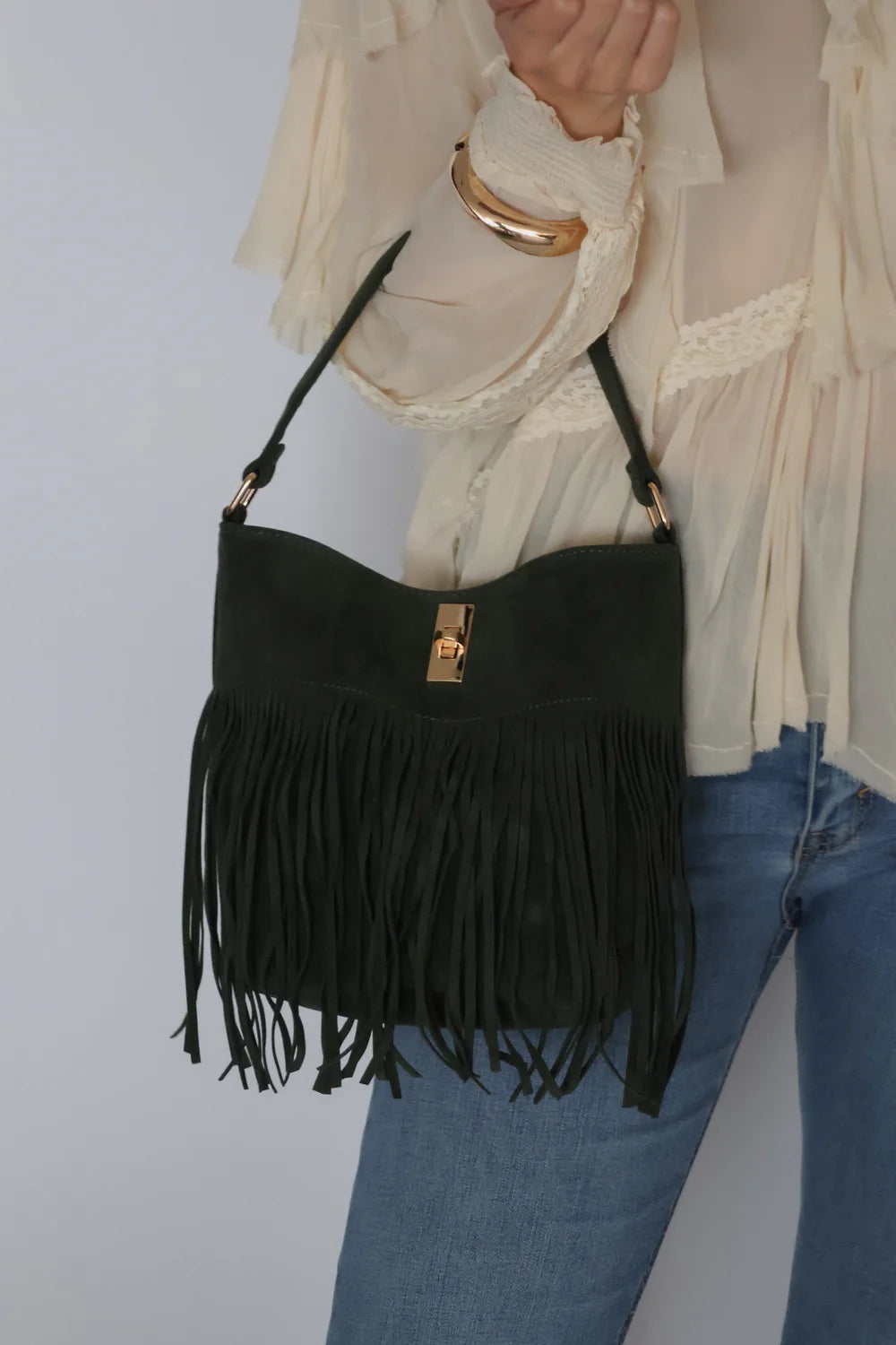 A dark green suede fringe shoulder bag with gold-tone hardware, shown being worn by a person in a cream blouse and blue jeans against a light gray background.