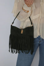 A dark green suede fringe shoulder bag with gold-tone hardware, shown being worn by a person in a cream blouse and blue jeans against a light gray background.