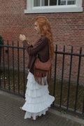 A woman with long red hair stands outdoors against a red brick wall and black metal fence, wearing a brown jacket, a long white ruffled skirt, and carrying a brown fringe shoulder bag.