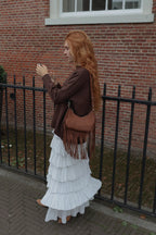 A woman with long red hair stands outdoors against a red brick wall and black metal fence, wearing a brown jacket, a long white ruffled skirt, and carrying a brown fringe shoulder bag.