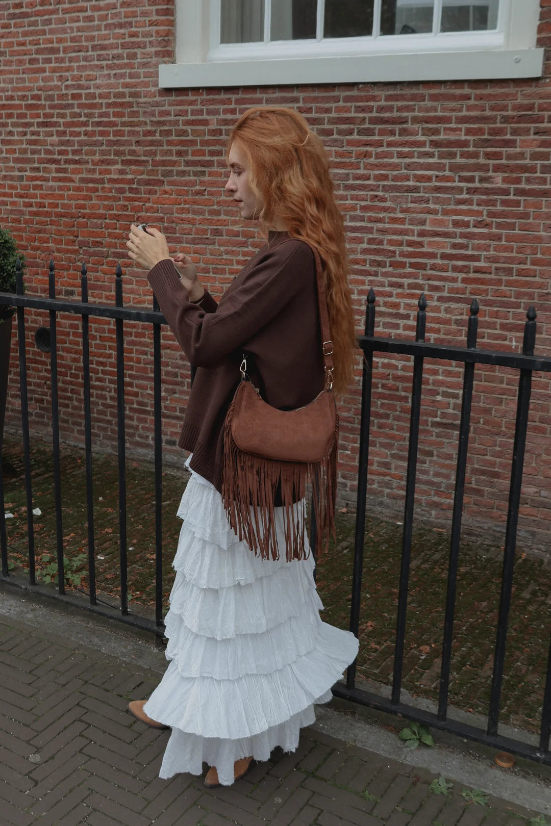 A woman with long red hair stands outdoors against a red brick wall and black metal fence, wearing a brown jacket, a long white ruffled skirt, and carrying a brown fringe shoulder bag.