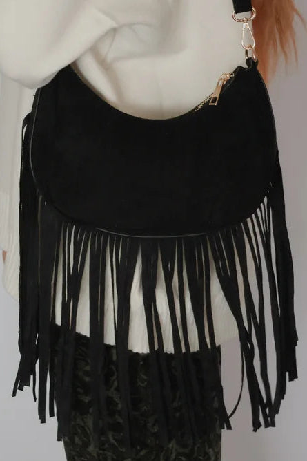 A black suede crescent moon shoulder bag with long fringe, worn crossbody over a cream-colored top.