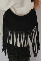 A black suede crescent moon shoulder bag with long fringe, worn crossbody over a cream-colored top.