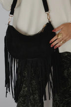 A black suede crescent-shaped shoulder bag with long fringe, worn by a model in a cream sweater and dark patterned pants against a light gray background.
