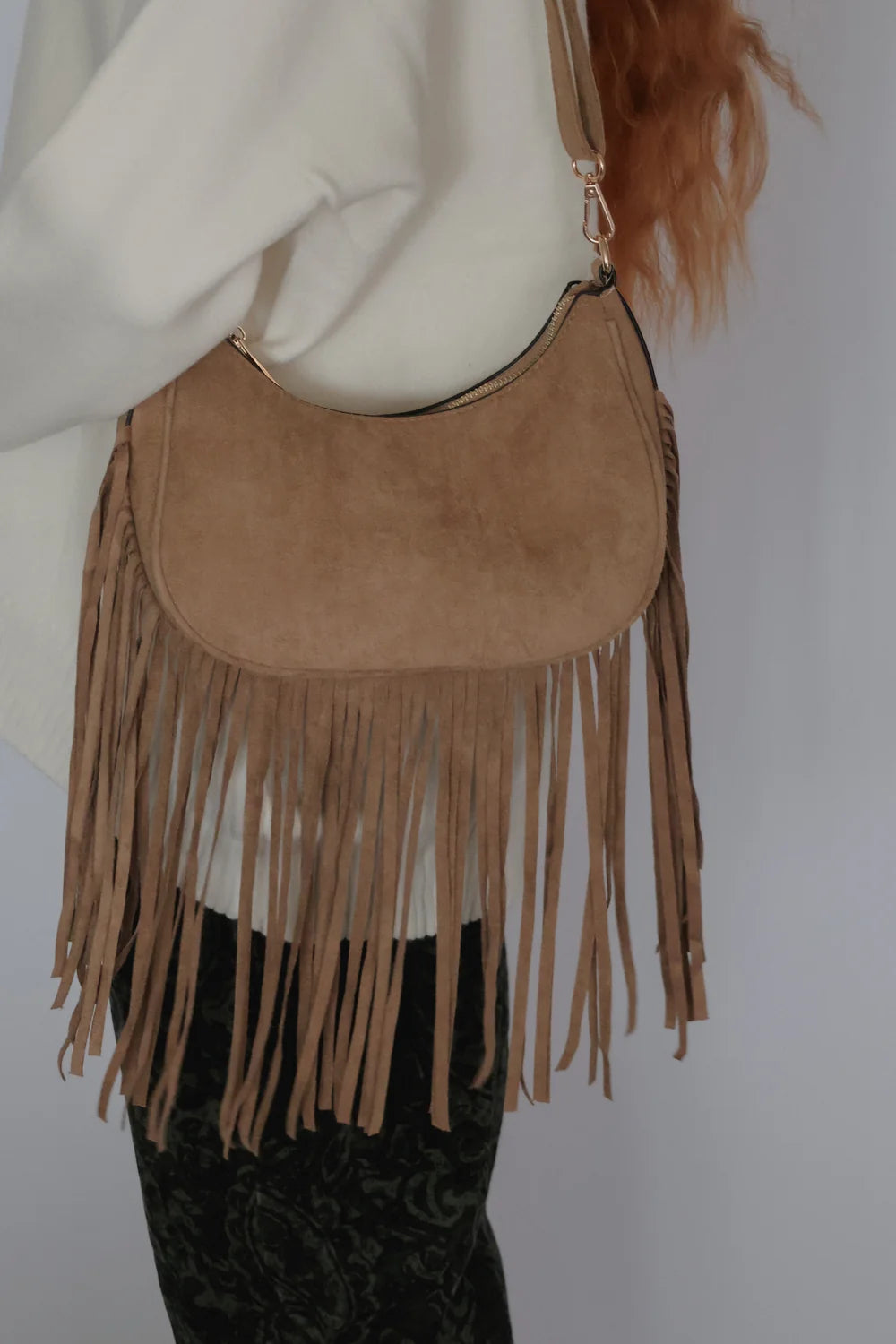 A tan suede crescent-shaped shoulder bag with long fringe, worn crossbody over a white top and dark patterned pants.