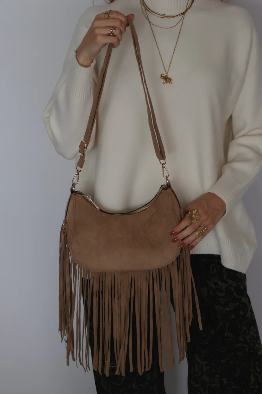 A person wearing a cream knit sweater and layered gold necklaces carries a tan suede crescent-shaped fringe shoulder bag against a plain light background.