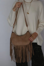 A person wearing a cream knit sweater and layered gold necklaces carries a tan suede crescent-shaped fringe shoulder bag against a plain light background.
