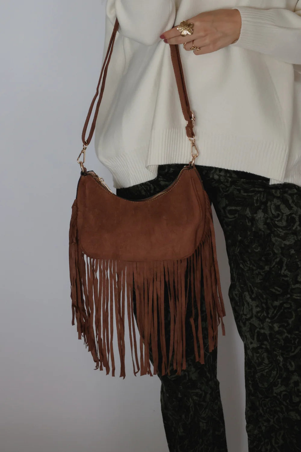 A brown suede crescent-shaped fringe handbag with a matching shoulder strap, worn crossbody by a person wearing a cream sweater and dark patterned pants.